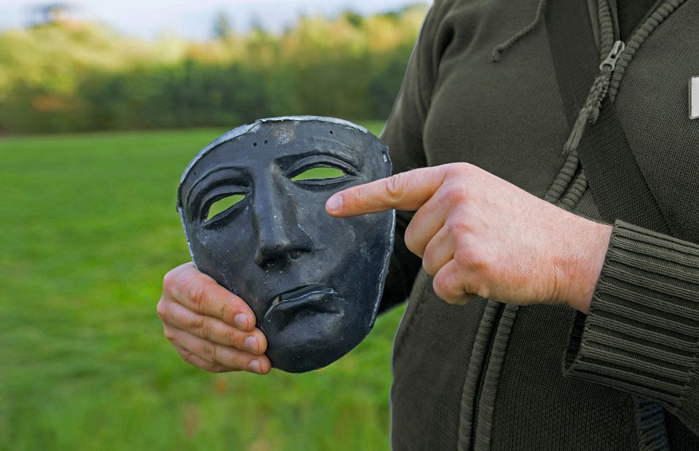 Mann zeigt auf Nachbildung der Reitermaske in seiner Hand.
