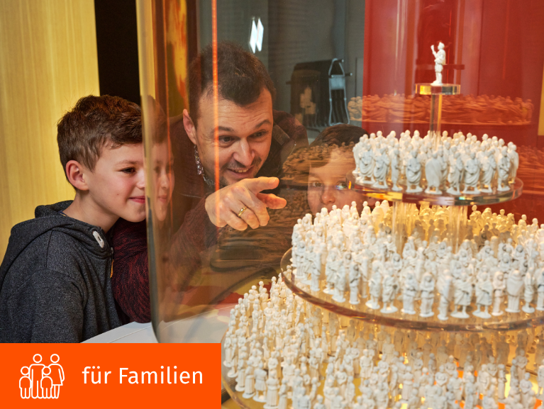 Mann zeigt mit Finger auf Miniaturlegionäre in Vitrine, Junge daneben lächelt.