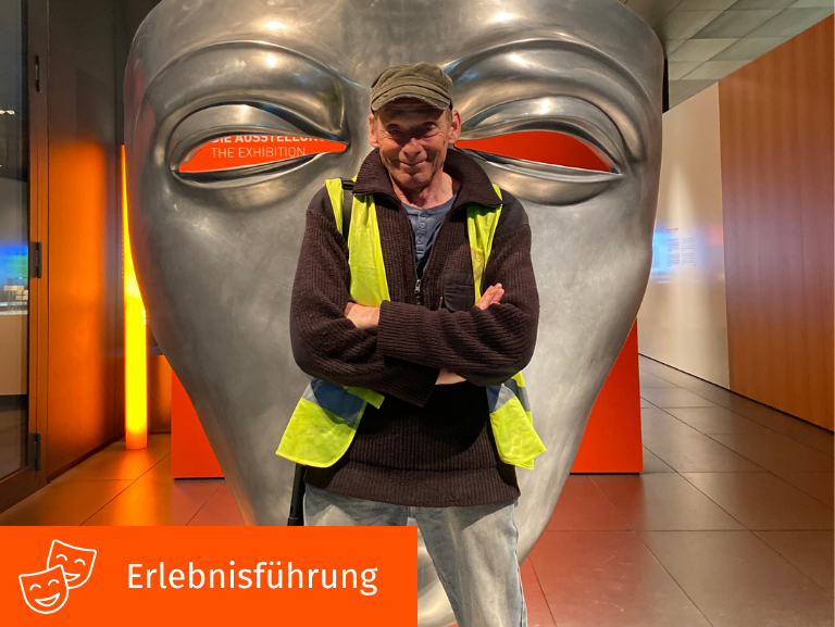 Mann steht lächelnd, mit verschränkten Armen vor großer Reitermaske.